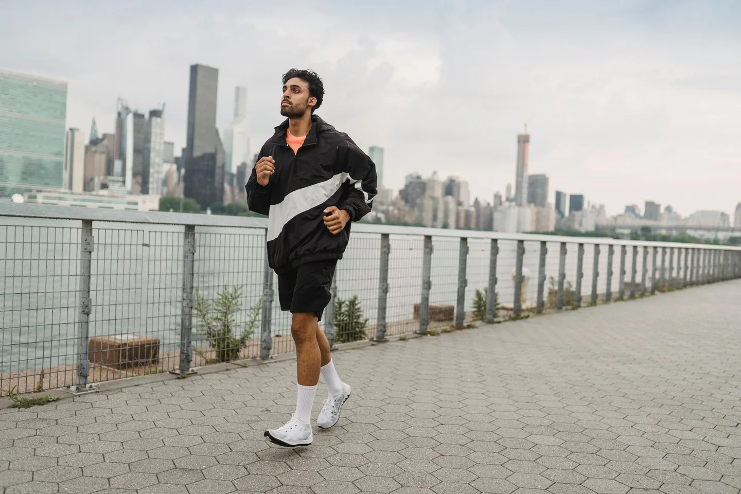 Candid urban lifestyle photograph of a runner in neutral sportswear and light knit running shoes moving along a riverside city path on an overcast morning