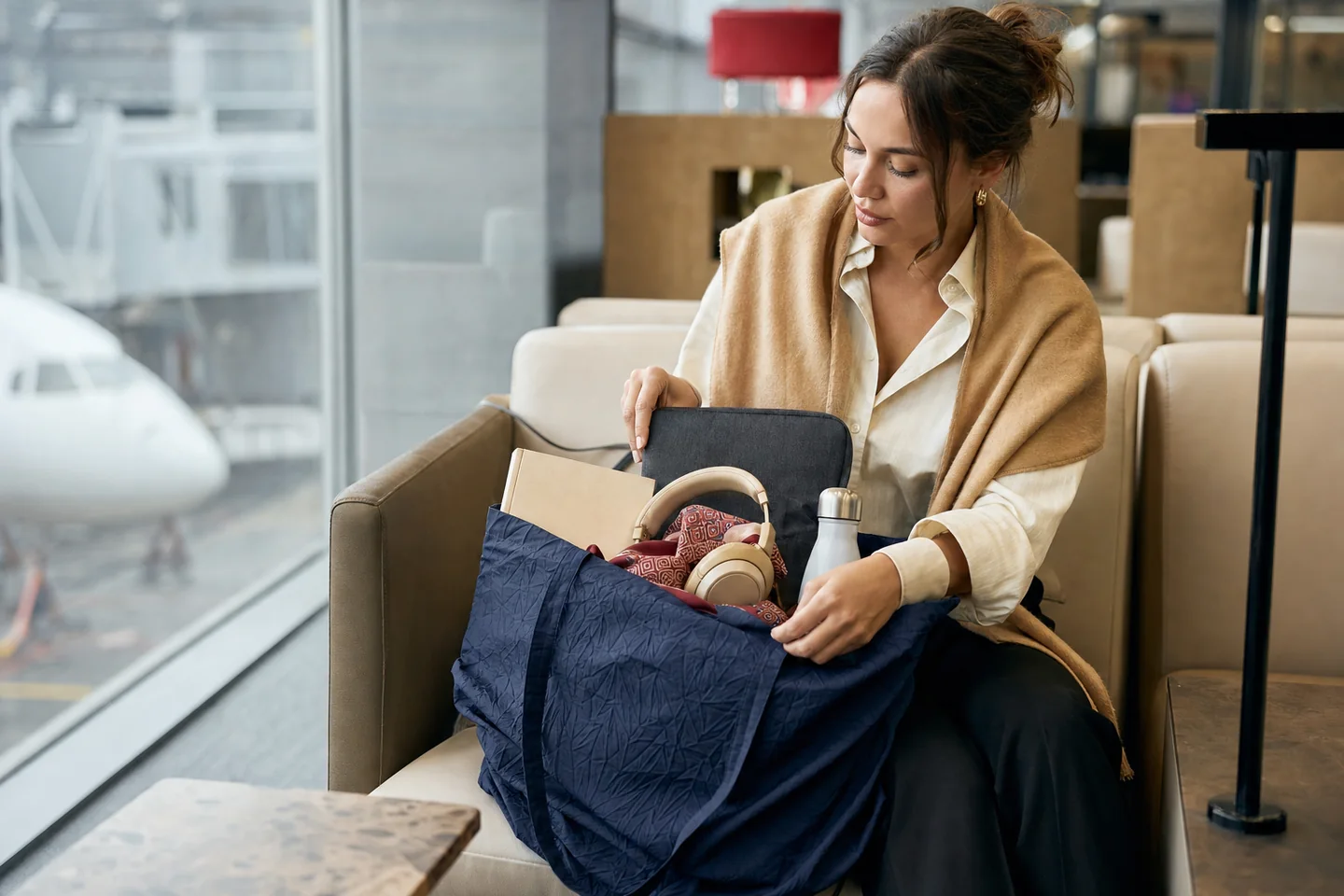 Natural airport lounge photograph of a traveler organizing a foldable nylon tote with book, scarf, headphones and a padded laptop sleeve